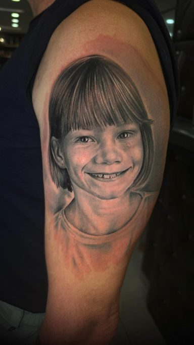 Porträt eines lächelnden Mädchens als Tattoo auf einem Oberarm. Tattoo von Anne Schwarte.